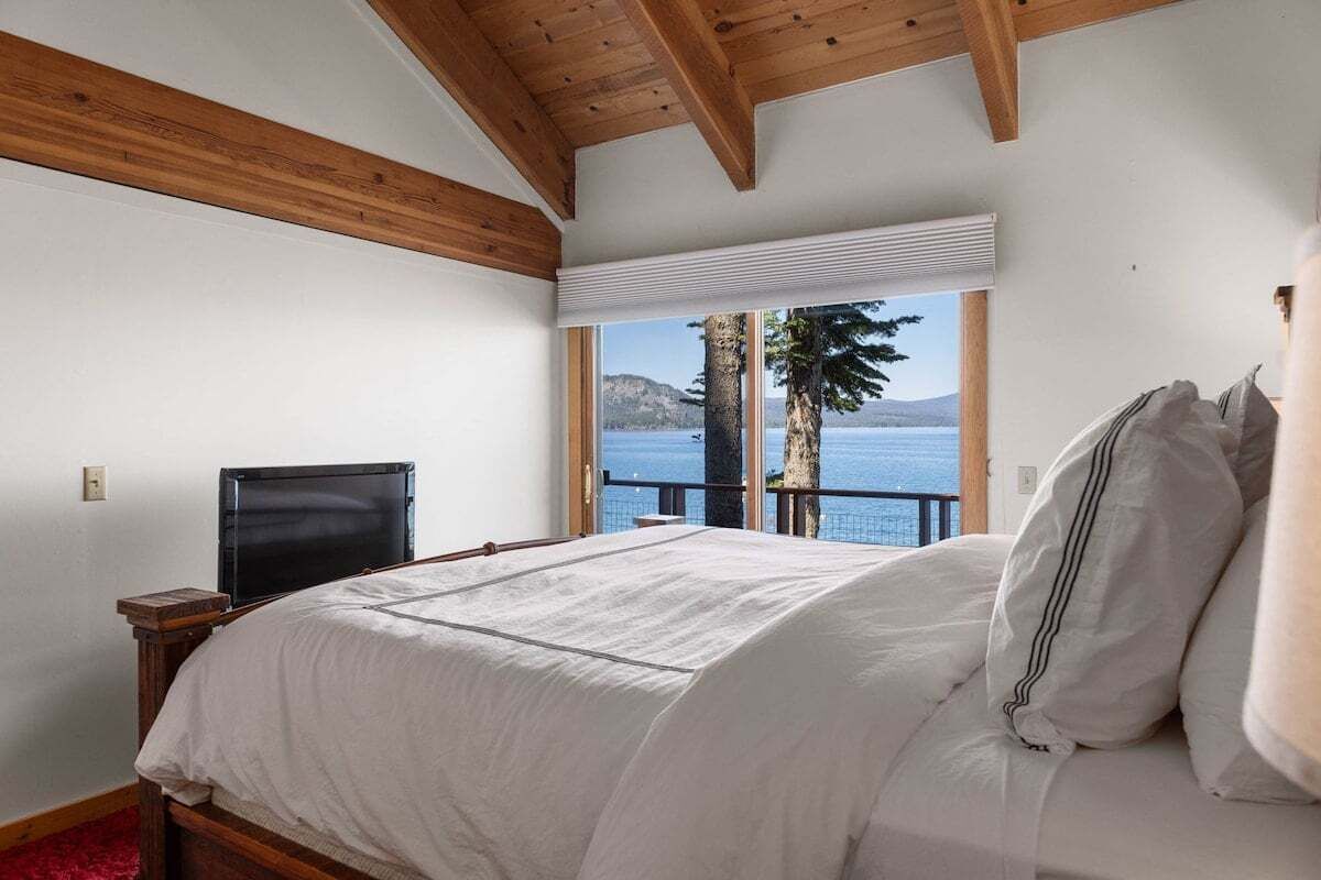 Une chambre avec un grand lit, des poutres en bois, une télévision et une fenêtre donnant sur un lac et des montagnes.