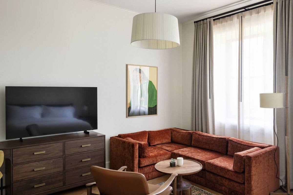 Moderni hotellihuone, jossa on kulmasohva, taulutelevisio, puinen sivupöytä ja taulu seinällä.