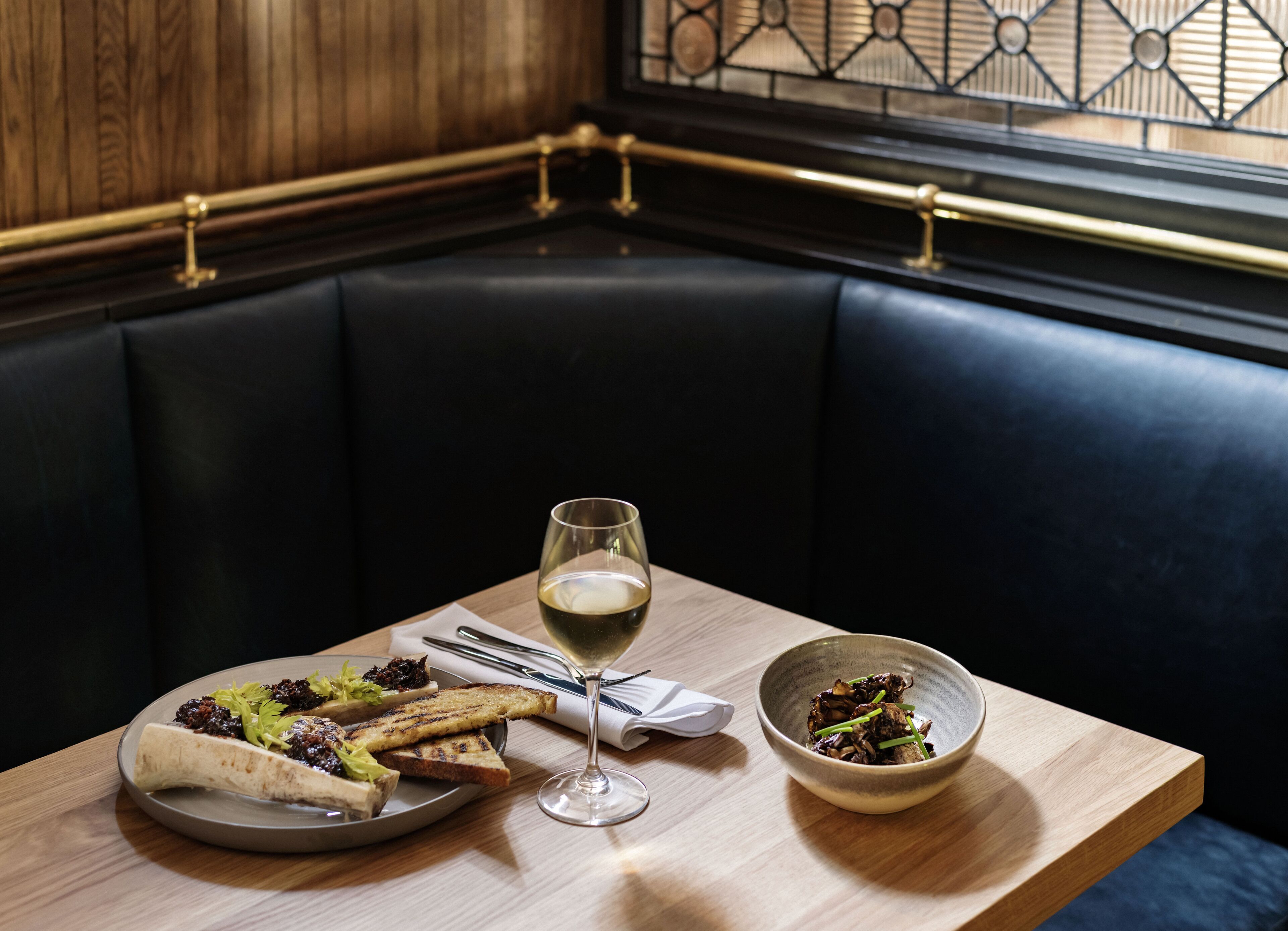Una mesa de comedor con un plato de comida, una copa de vino y un bol de ensalada en una zona de estar con paredes de madera y barandilla de metal.