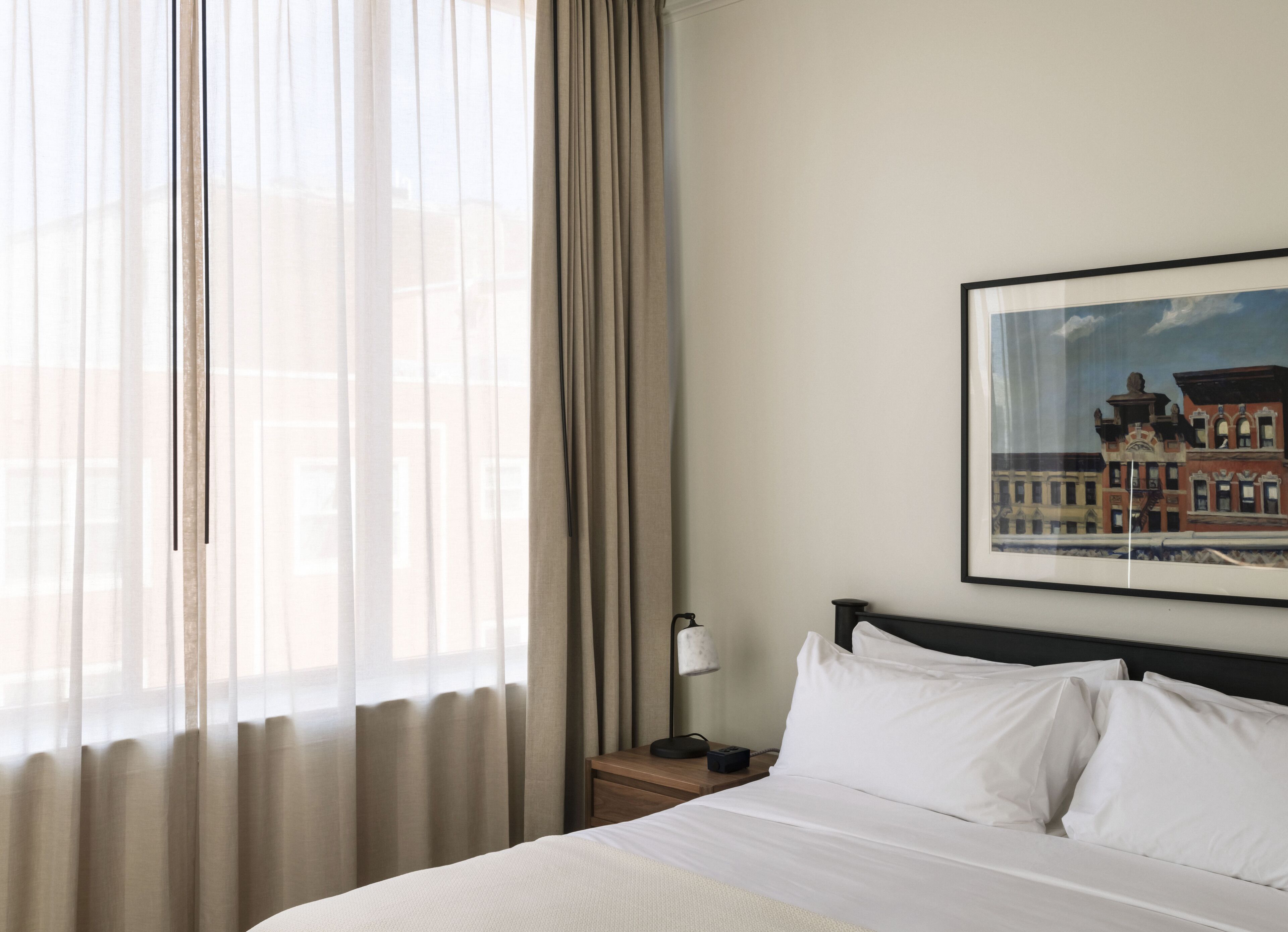 Une chambre d’hôtel avec un lit, une table de chevet, une lampe et une fenêtre avec des rideaux légers.