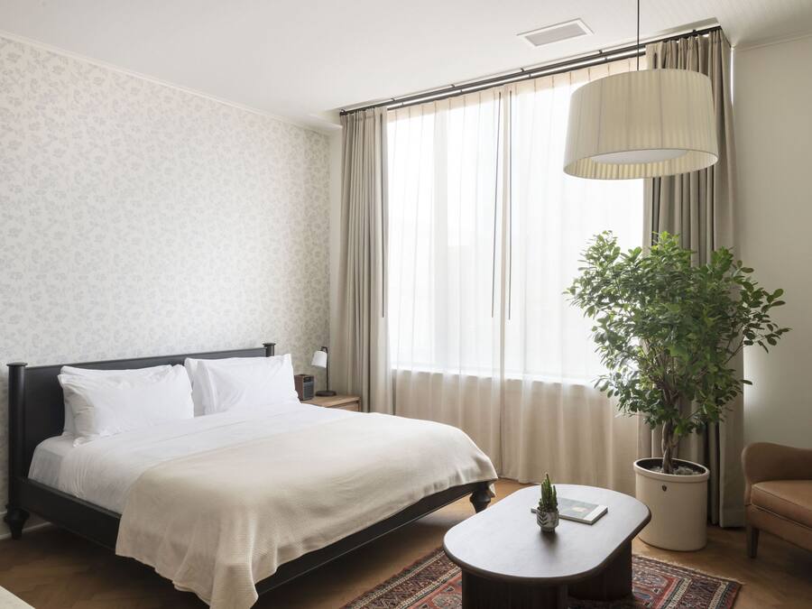 Een hotelkamer met een bed, een nachtkastje met een lamp, een potplant, een bank en een groot raam met gordijnen.