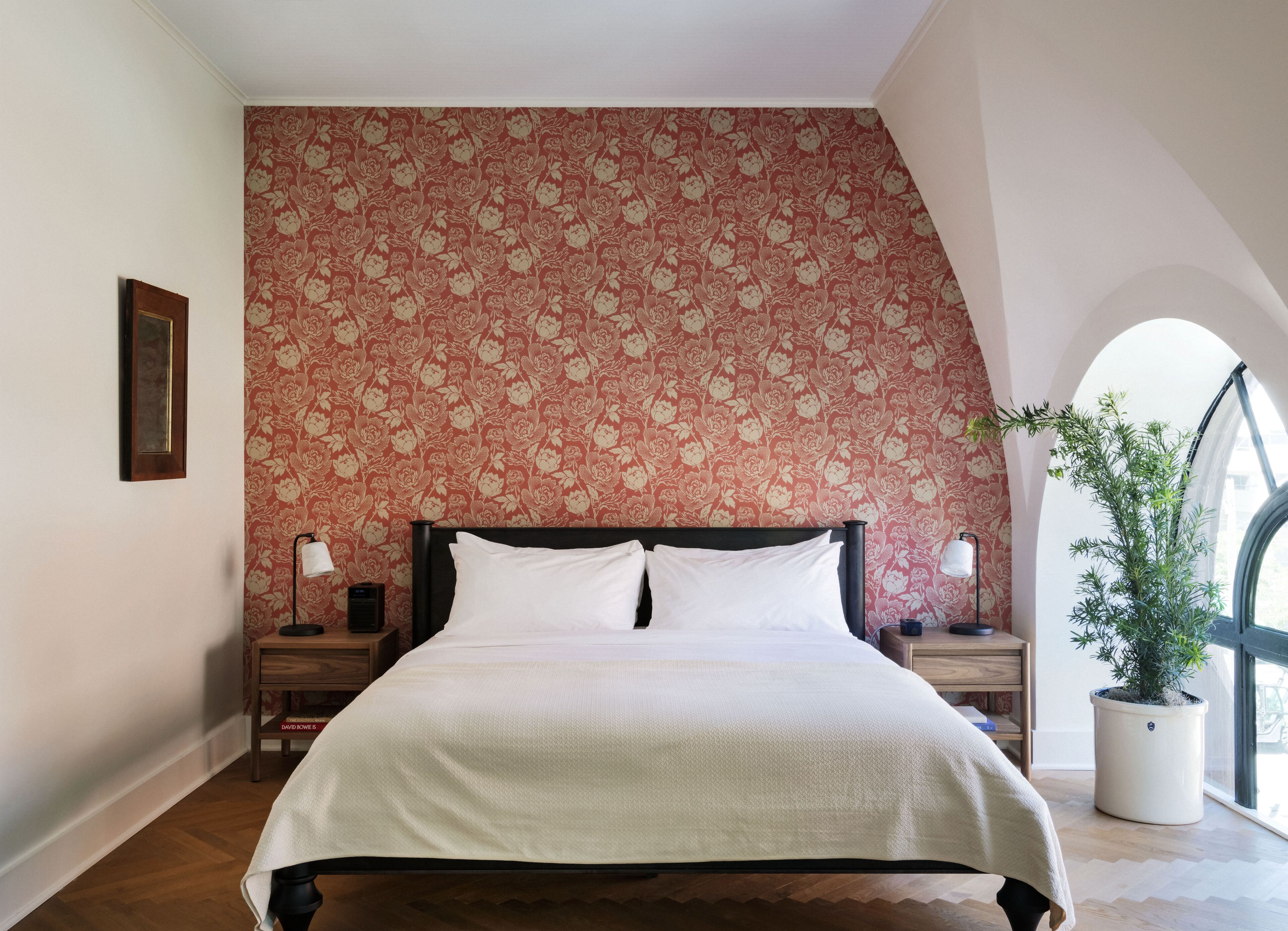 A bedroom with a floral wallpaper, a bed with white linens, two bedside tables with lamps, and a potted plant by the window.