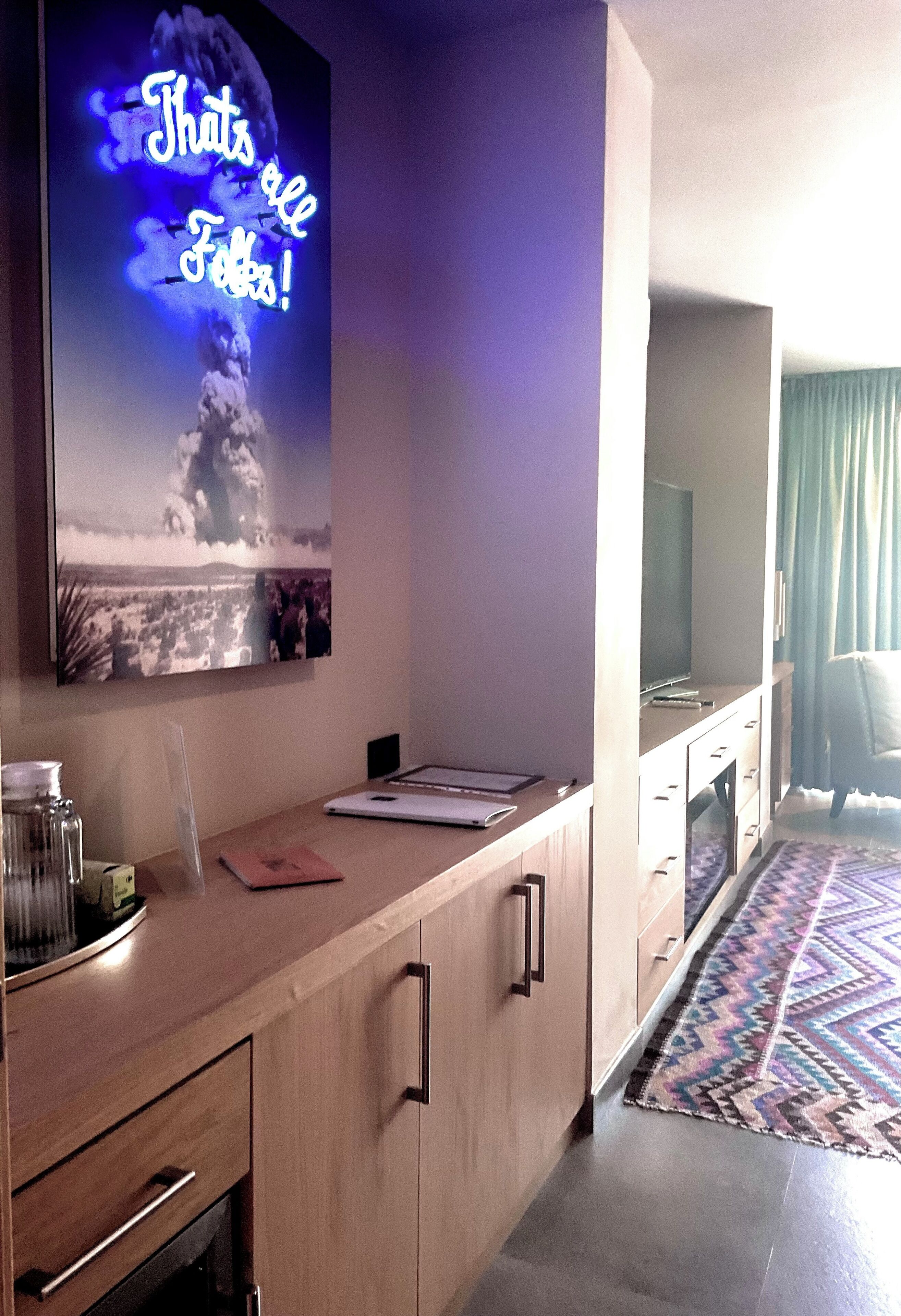 Une chambre d’hôtel moderne avec un bureau en bois, un téléviseur à écran plat, une grande affiche et un tapis à motifs.