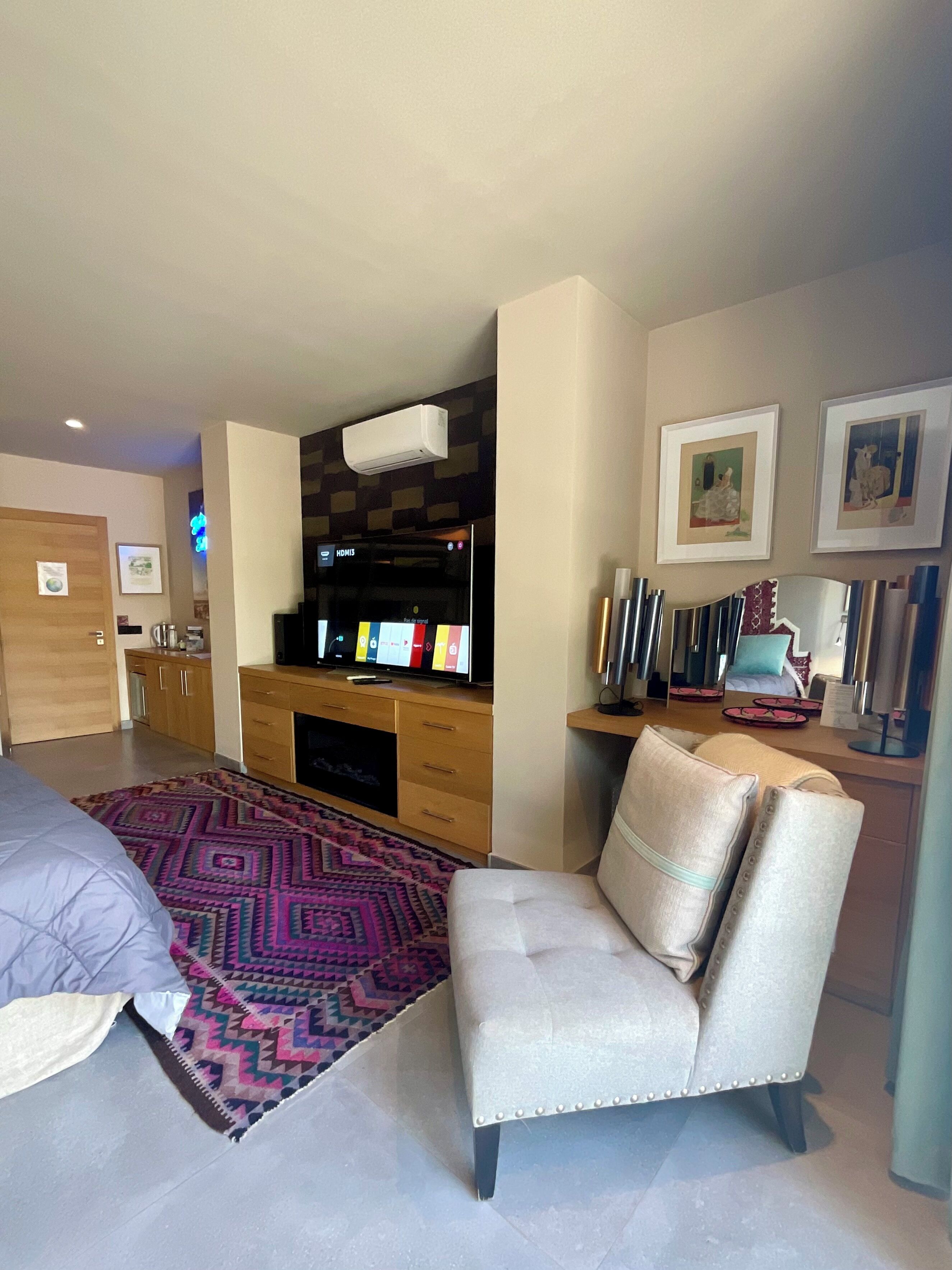 A modern bedroom with a bed, a seating area with a chair, a desk with a computer, and a TV.