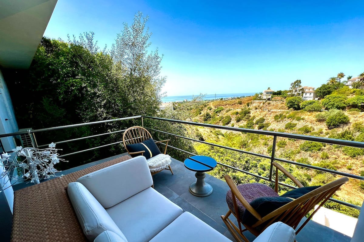 Un balcón con un sofá blanco, una silla de madera y una pequeña mesa. El balcón ofrece vistas a un paisaje montañoso con vegetación y un cielo despejado.