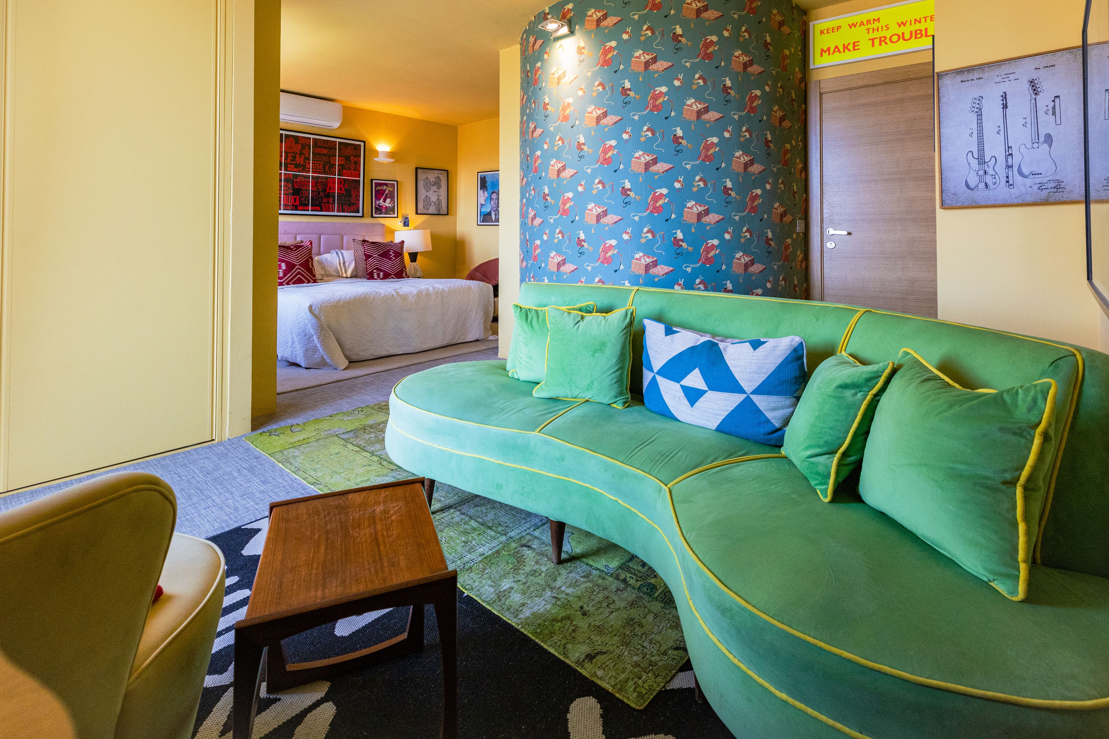 A room with a green sofa, a wooden chair, a bed with red and white pillows, a patterned wall, and a framed patent drawing.