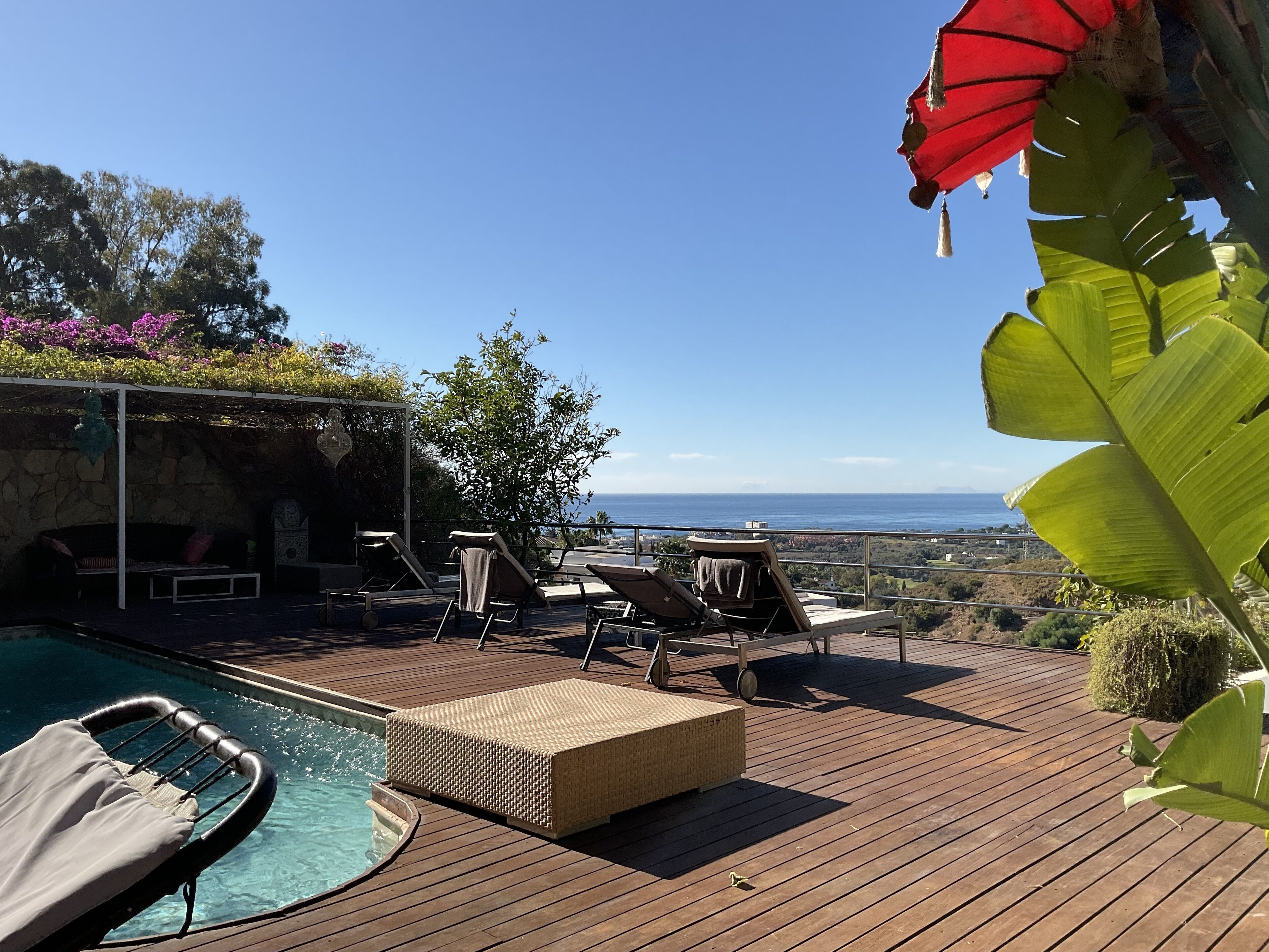 Une terrasse en bois avec des chaises longues et une piscine, offrant une vue panoramique sur l’océan et les collines.