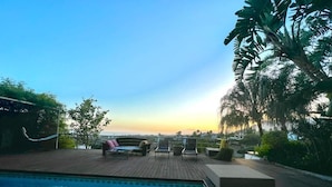 Zona de piscina con terraza de madera, sillones de descanso y vistas a la puesta de sol sobre el océano.