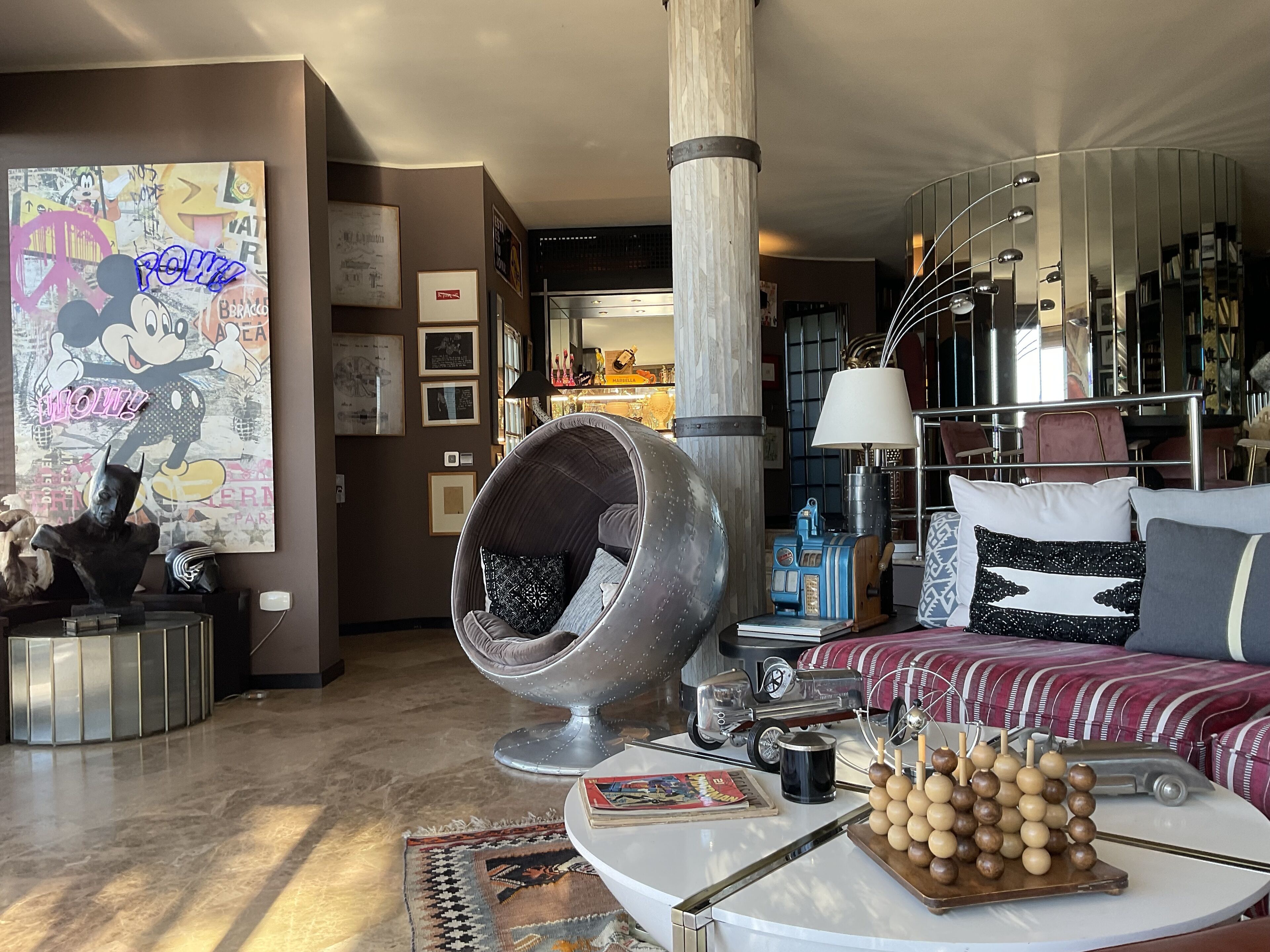 Una sala moderna con un sillón grande y redondo de metal, una mesa de centro redonda con un juego de mesa, un sofá con cojines estampados y un gran cartel de Mickey Mouse en la pared.