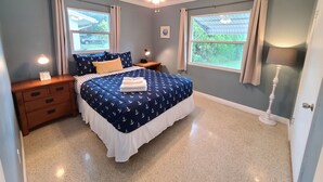 A bedroom with a navy blue bed, white curtains, a ceiling fan, and a window with a view of greenery.