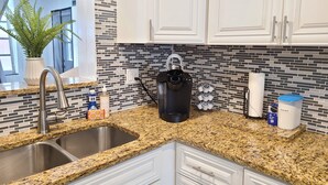 A kitchen with granite countertops, a stainless steel sink, white cabinets, a coffee maker, and a potted plant.