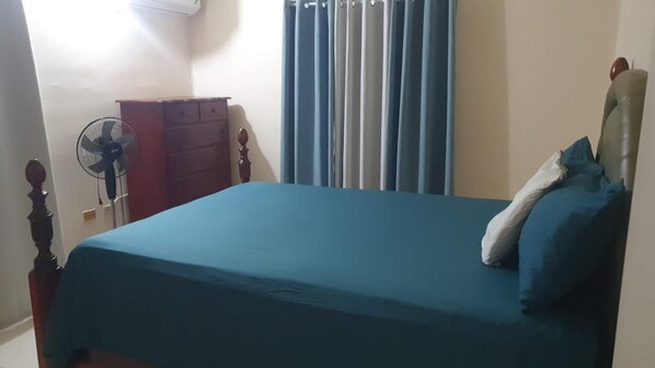 Een slaapkamer met een bed, een dressoir, een ventilator en een raam met gordijnen.