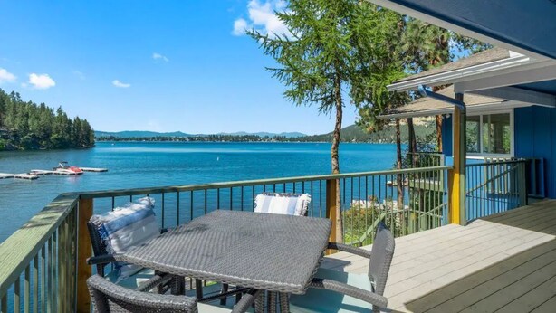 Una terraza de madera con una mesa y sillas da a un lago, con vistas claras del agua y colinas distantes.