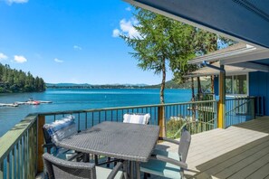A wooden deck with a table and chairs overlooking a lake, with a clear view of the water and distant hills. - Incredible Waterfront Lakehouse on Liberty Lake (Liberty Lake)