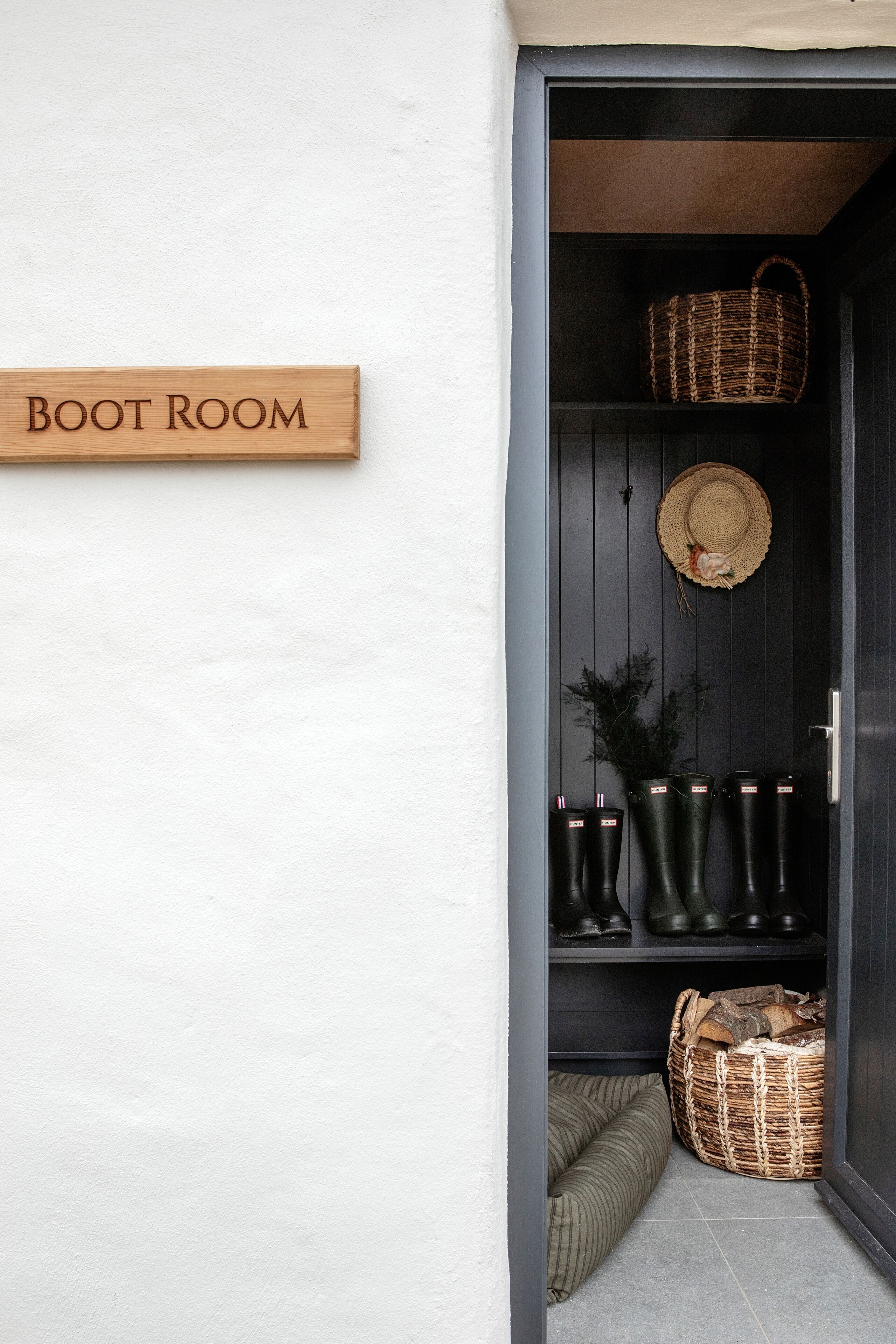 A dark wooden door with a sign reading "BOOT ROOM" above it. Inside, there are wicker baskets, a woven hat, and several pairs of black boots.