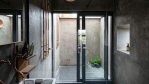 Un bany modern amb una porta de vidre que dóna a un pati, elements de bronze i una petita estant amb productes de tocador.