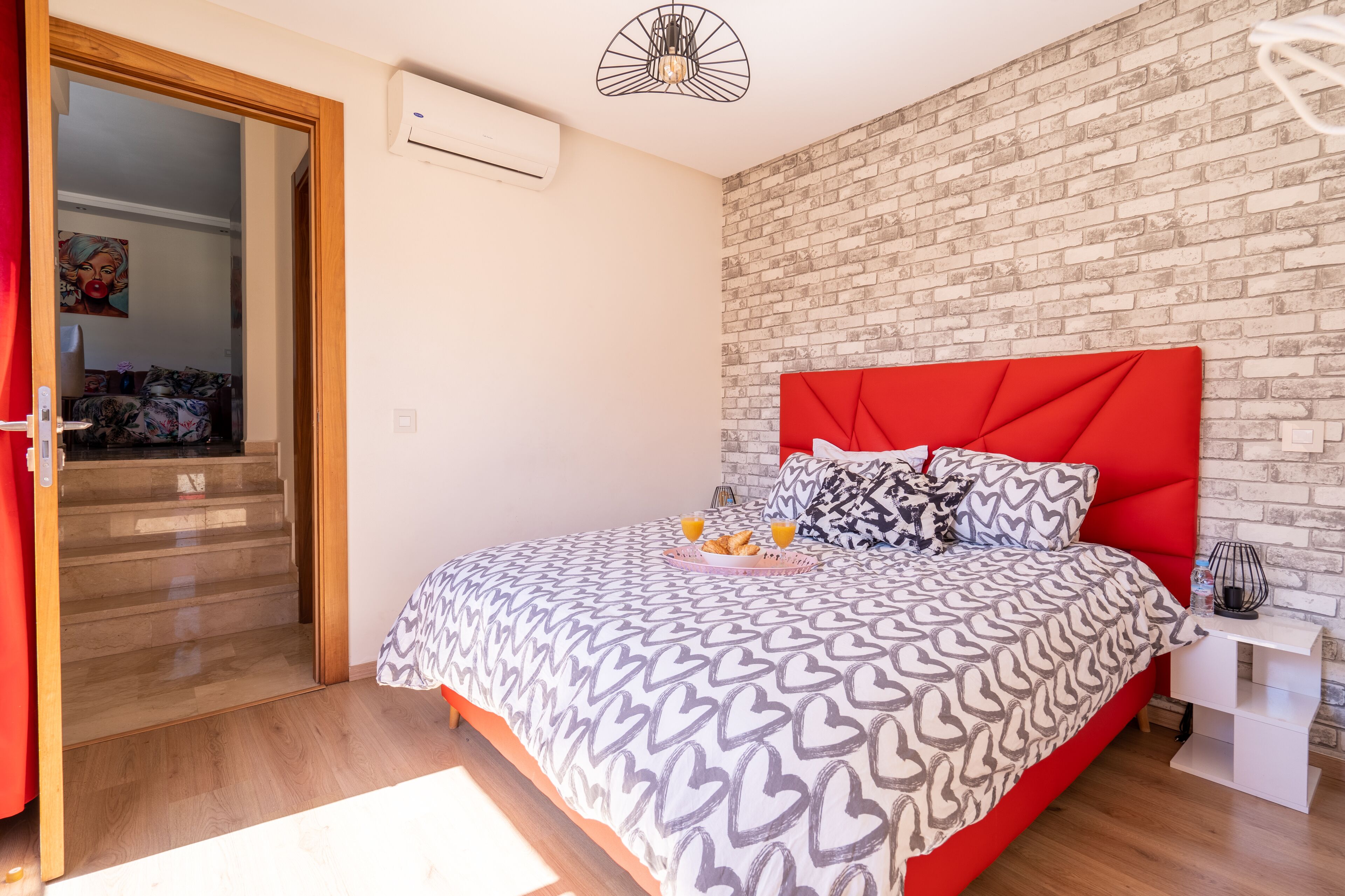 Une chambre à coucher avec une tête de lit rembourrée de tissu rouge, une couvre-lit à motifs et un plateau contenant des croissants et du jus d’orange.