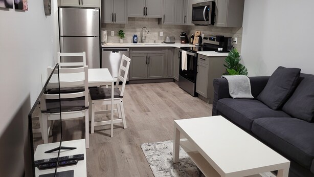 Cocina moderna con gabinetes grises, un refrigerador de acero inoxidable, un microondas y un lavavajillas. Hay un comedor con sillas y un sofá gris en la sala.