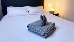 Una cama bien hecha con sábanas y almohadas blancas, un cabecero oscuro y una mesita de noche con una lámpara y una caja de pañuelos.
