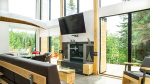 A modern living room with a large TV, a fireplace, and a view of the forest. - Le Baluchon Éco-villégiature (Saint-Paulin)