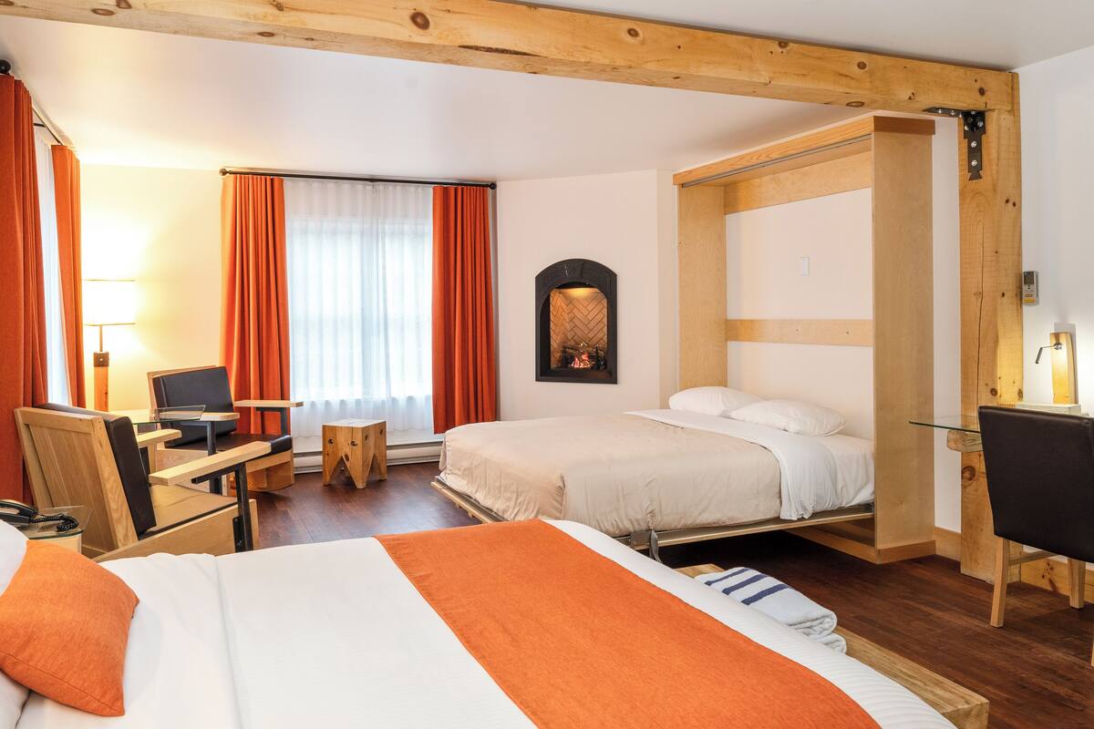 A hotel room with a bed, a desk, a chair, a fireplace, and a window with curtains.