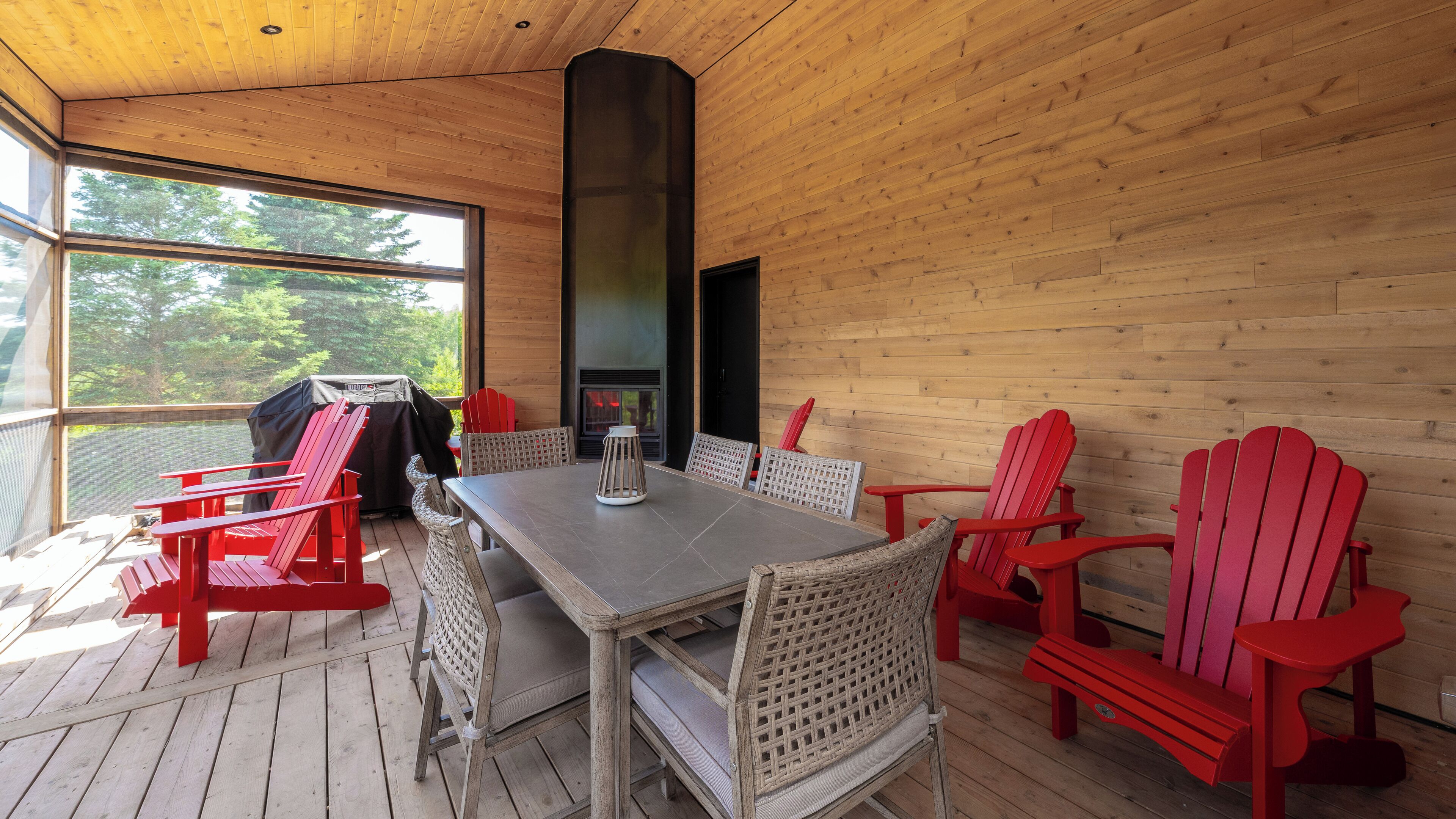 A wooden cabin with a dining area featuring red Adirondack chairs, a table, and a grill.
