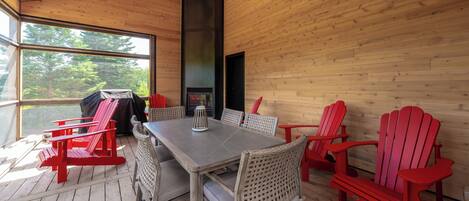 A wooden cabin with a dining area featuring red Adirondack chairs, a table, and a grill.