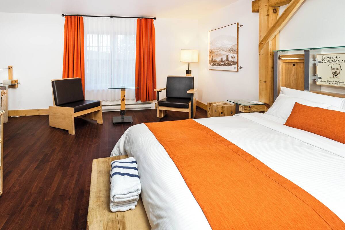 A hotel room with a large bed, two chairs, a desk, a small table, and a window with orange curtains.