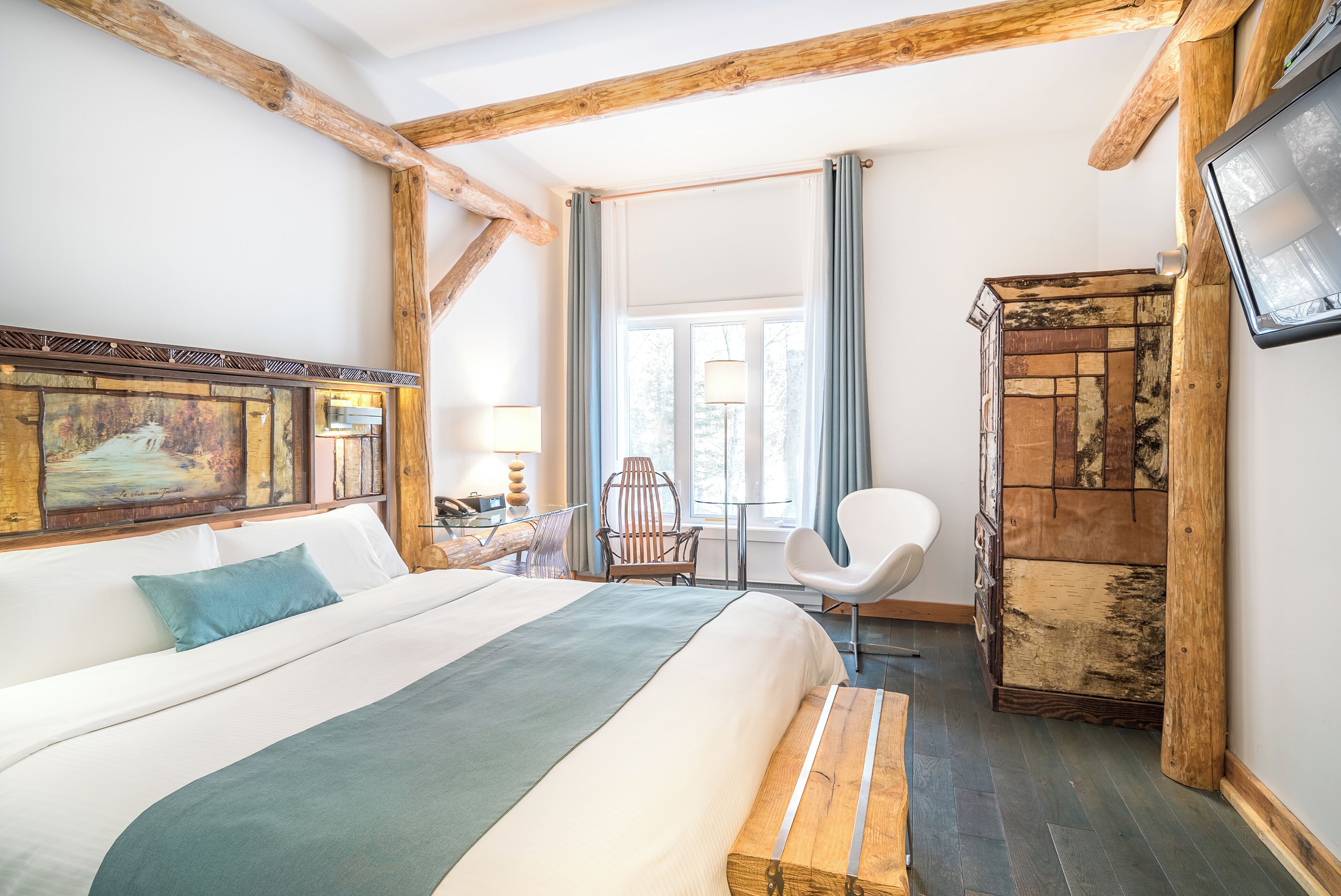 A hotel room with a large bed, wooden beams, a desk with a chair, a wardrobe, and a window with curtains.