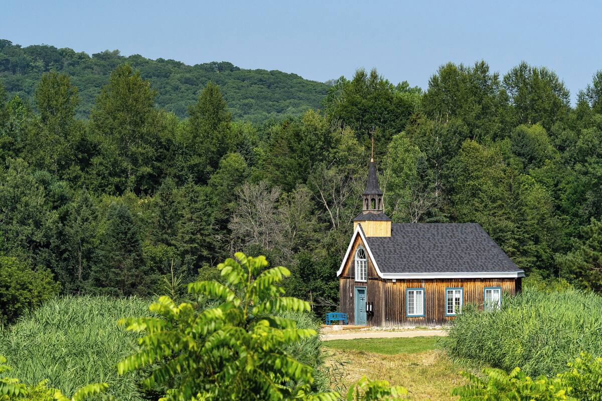 A wooden cabin with a steep roof and a bell tower, surrounded by dense forest and hills.