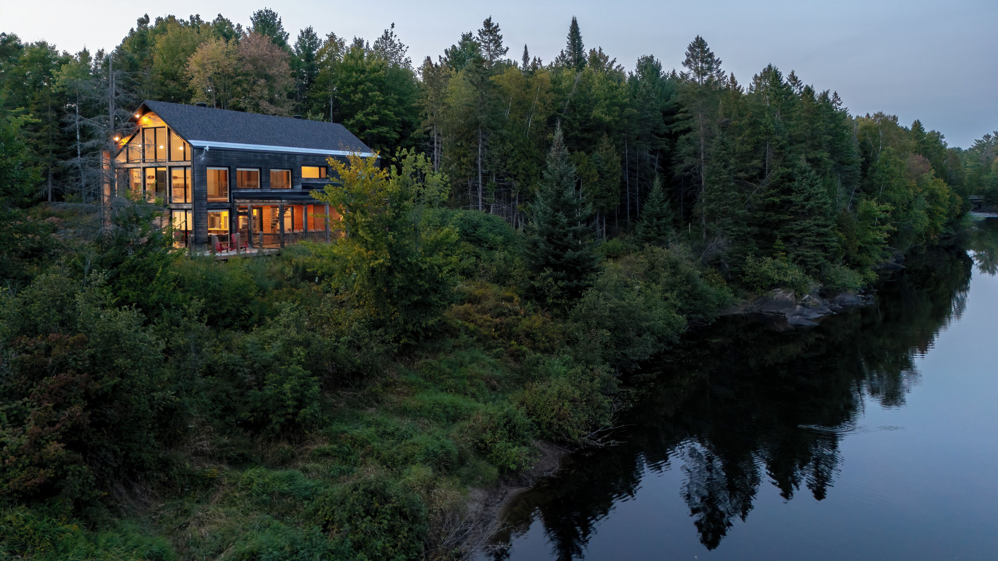 Un chalet au bord du lac, doté d’une terrasse et de grandes fenêtres, entouré d’une forêt dense et dont l’image se reflète sur l’eau.