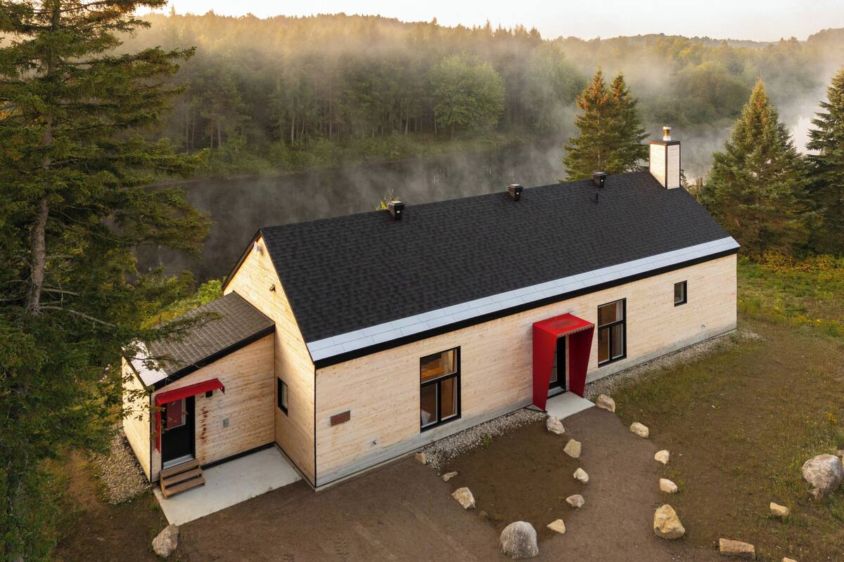 A cabin with a sloped roof, a chimney, and a red door. It is surrounded by trees and a forested area.