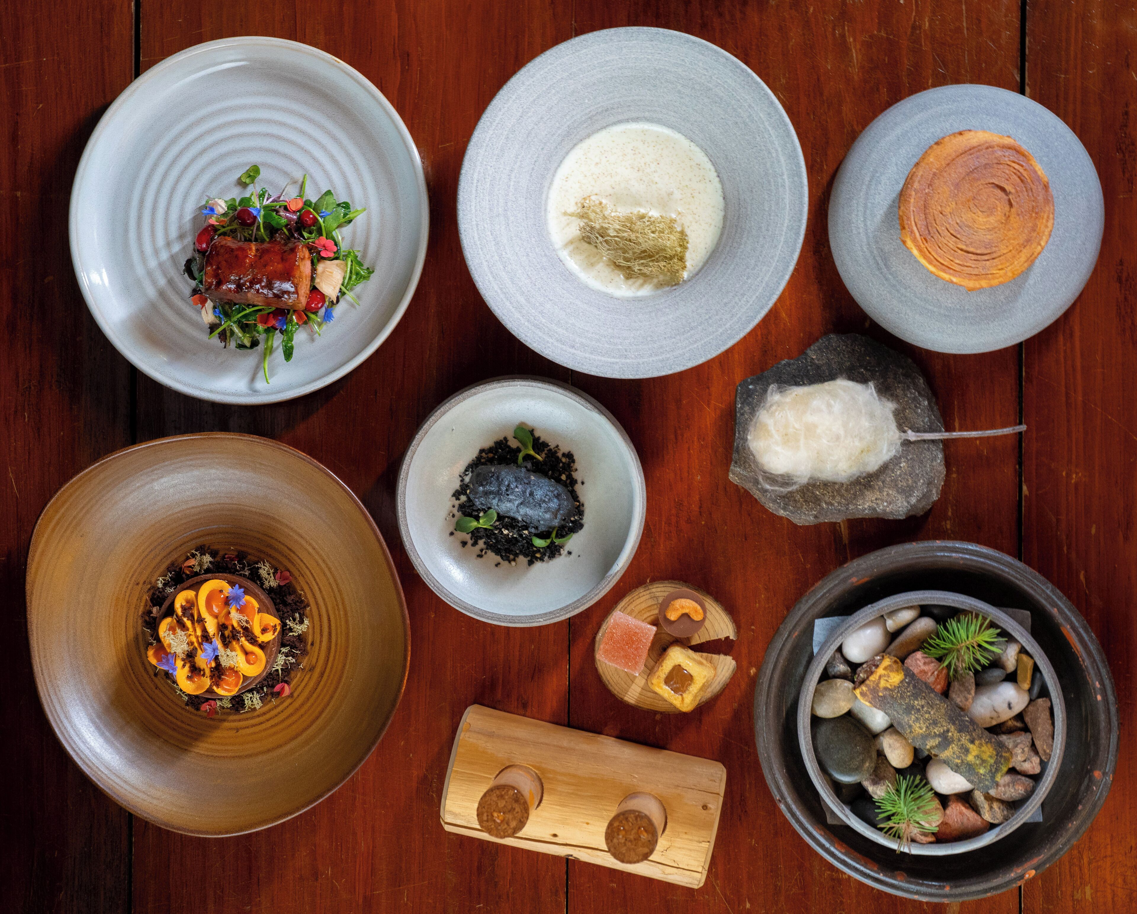 A variety of dishes on a wooden table, including a bowl of soup, a plate of meat, and a bowl of rice.