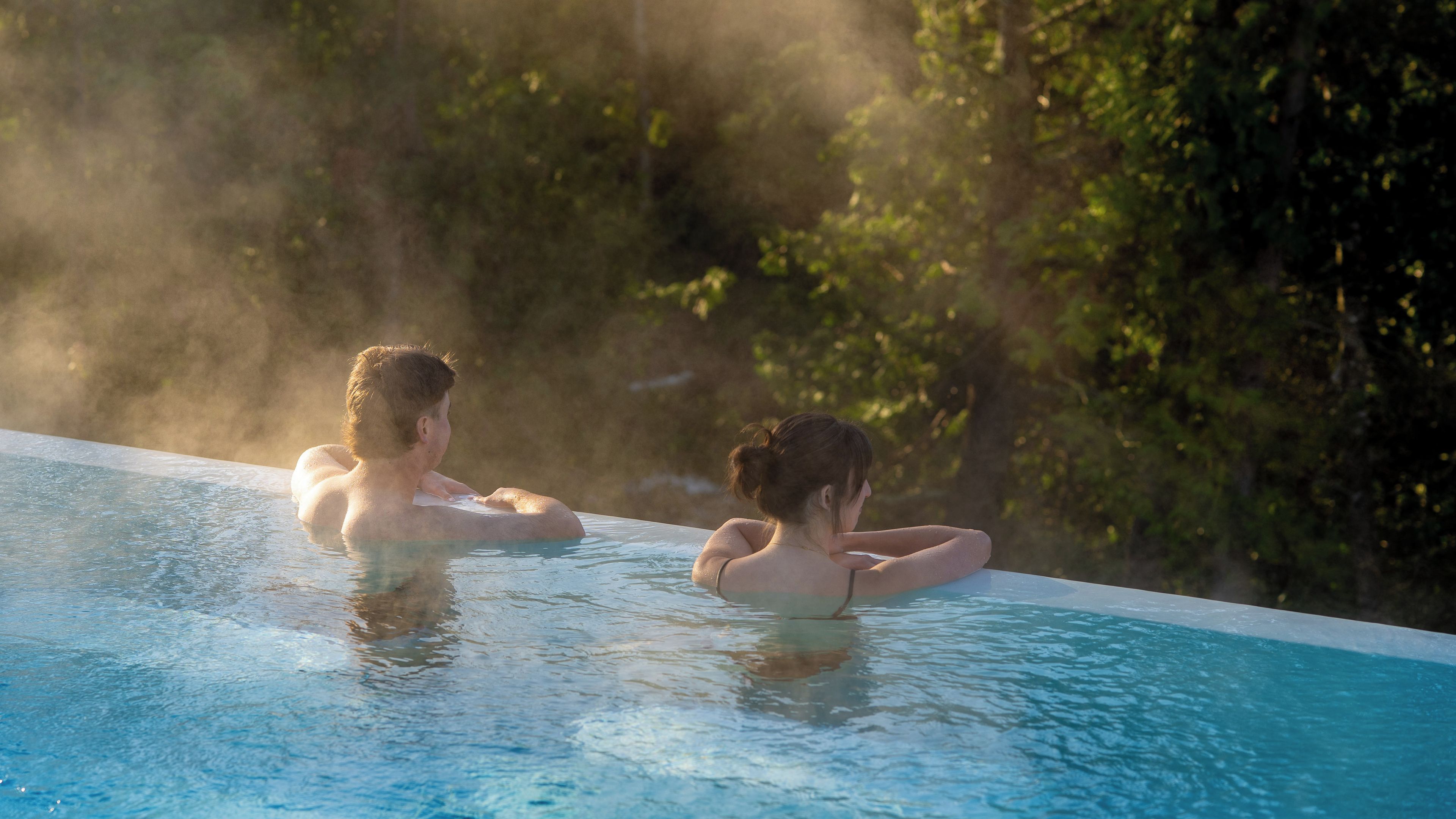 Two people in a swimming pool with steam rising from the water, surrounded by greenery.