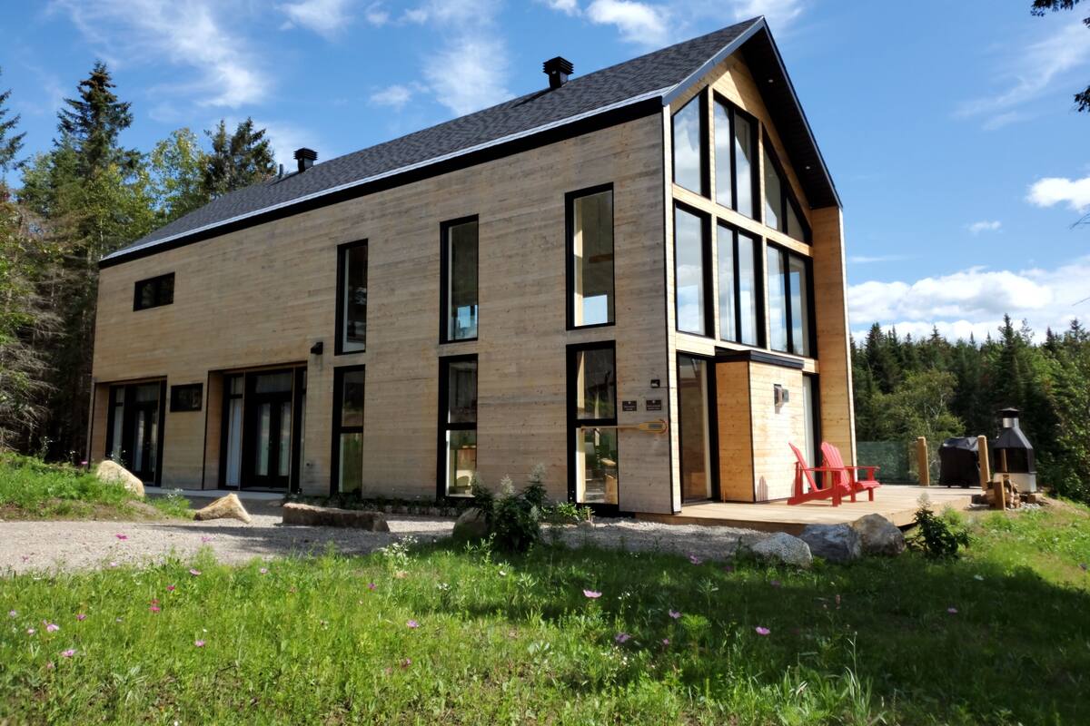 A modern two-story house with large windows, a wooden exterior, and a deck with outdoor seating.