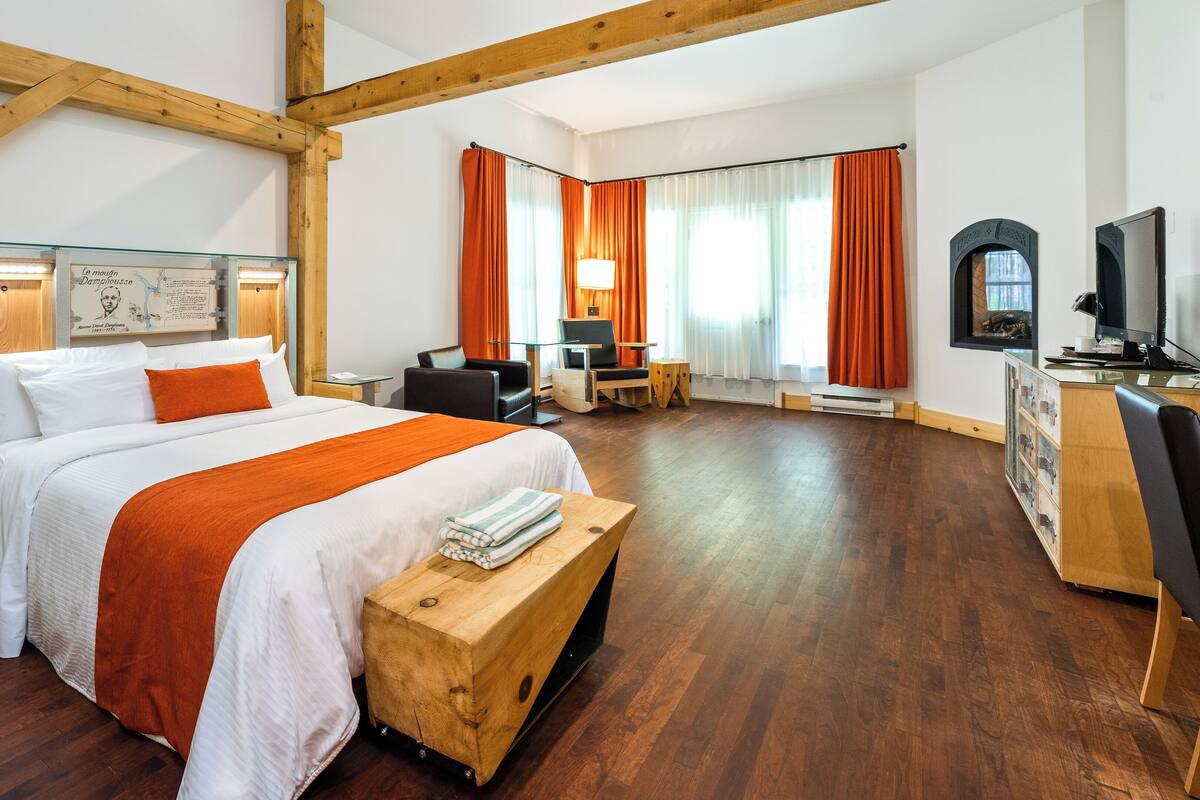 A hotel room with a large bed, wooden beams, a desk with a computer, a TV, and a window with curtains.