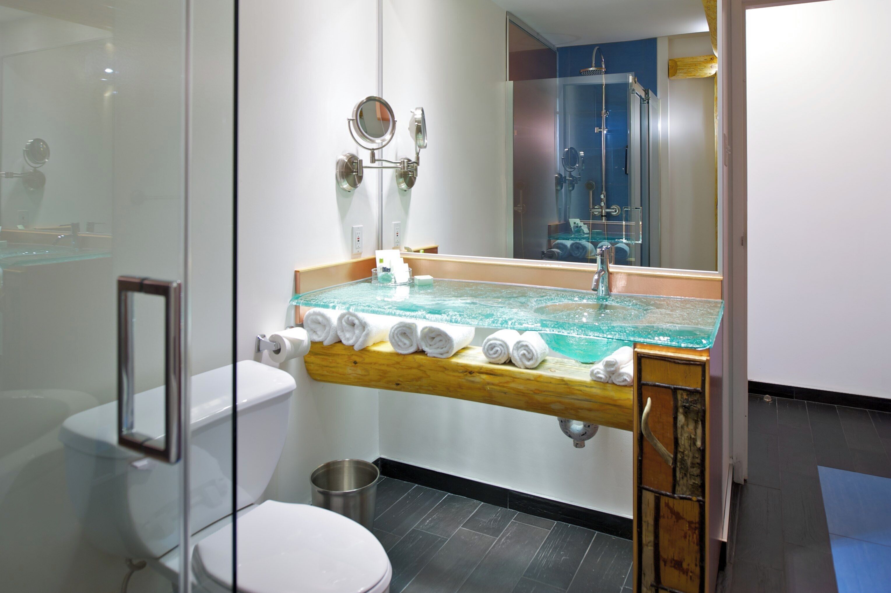 A modern bathroom with a glass-topped sink, a wooden shelf with towels, a toilet, and a glass shower enclosure.