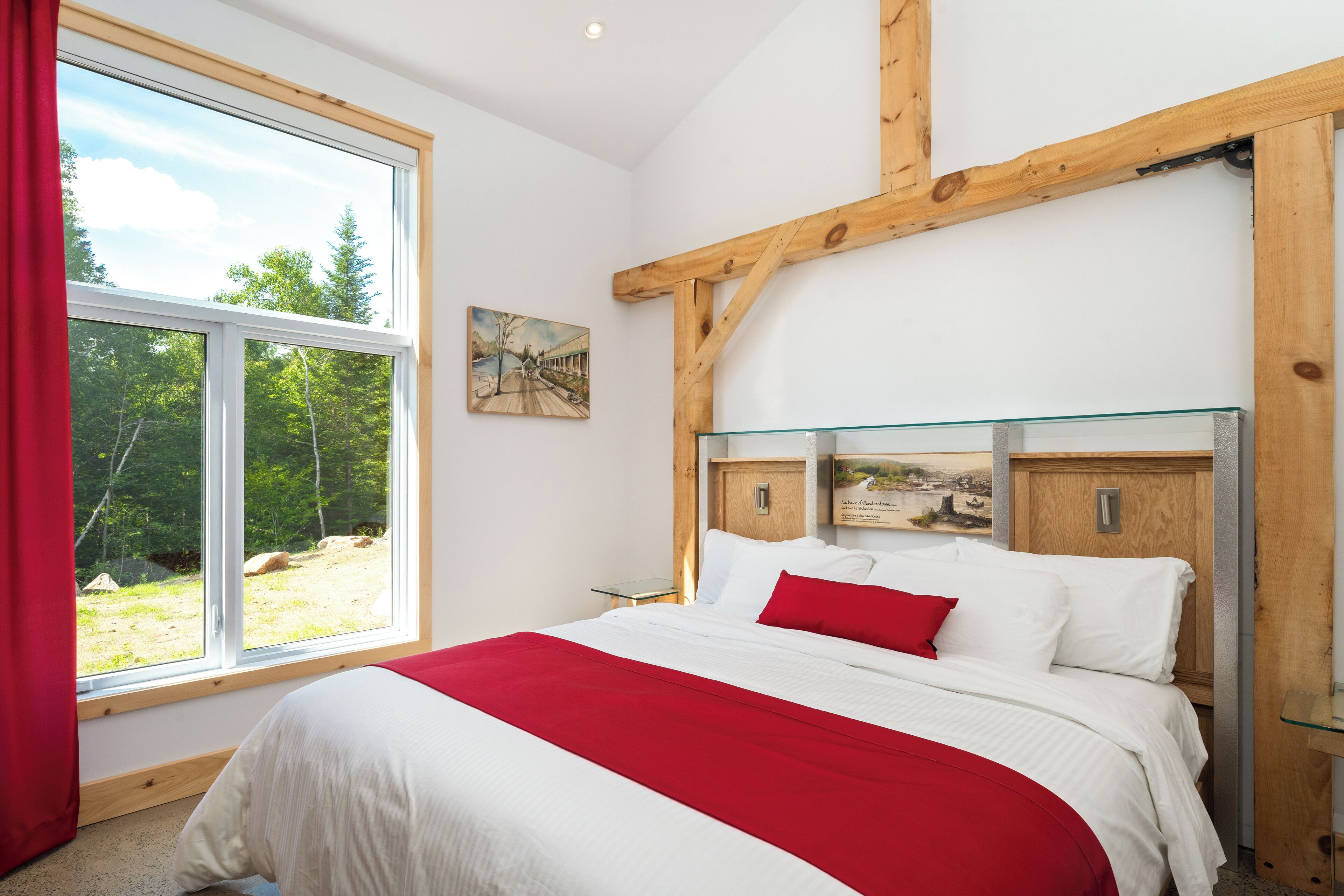 Un dormitorio con un ventanal grande, una cama con ropa de cama blanca y roja, y una estructura de techo de madera.