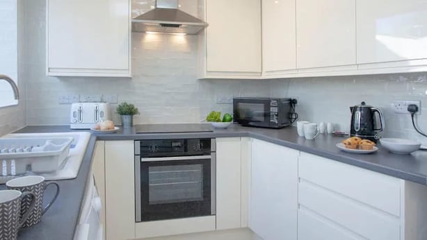 A modern kitchen with white cabinets, a stainless steel range hood, a built-in oven, a microwave, and a coffee maker.