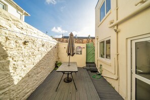 En smal udendørs terrasse med et bord og stole, et grønt jerngitter og en stenvæg.