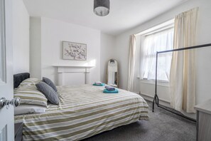 A hotel room with a bed, a mirror, a window with curtains, a radiator, and a wall-mounted picture. - Torquay 3-bed House, Sleeps 6, Pets Ok & Parking (Torquay)