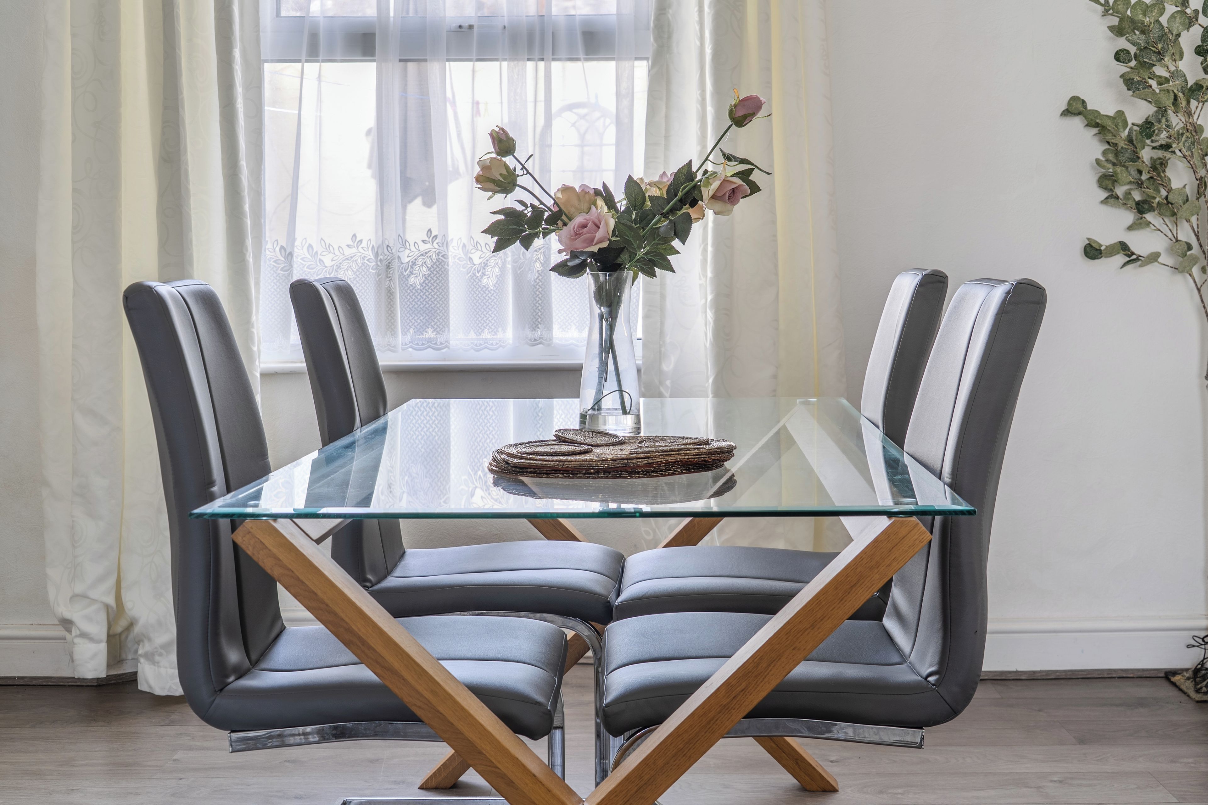 A modern dining table with a glass top and wooden legs, surrounded by four grey upholstered chairs.