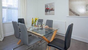 A dining area with a glass table, four chairs, a bottle of wine, a vase with flowers, and a painting on the wall.