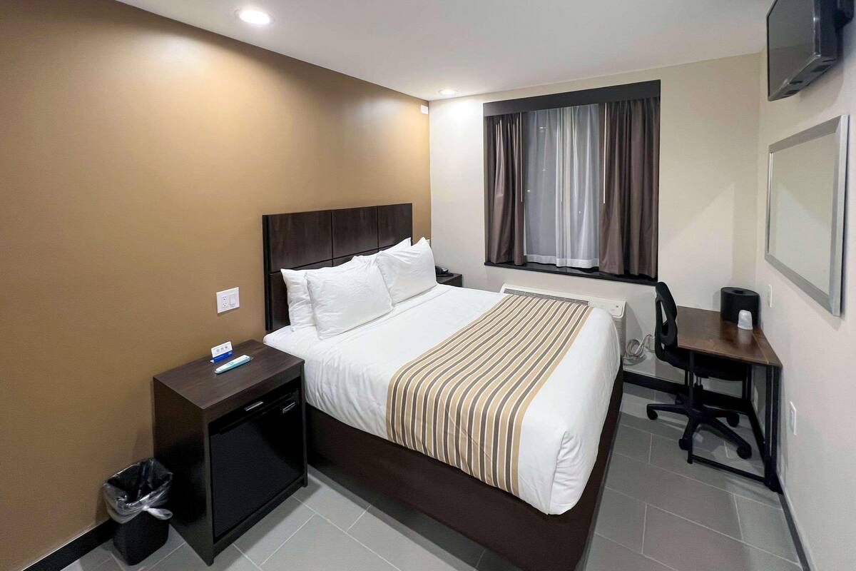 Una habitación de hotel con una cama grande, un escritorio, una silla, un televisor y un ventanal con cortinas.