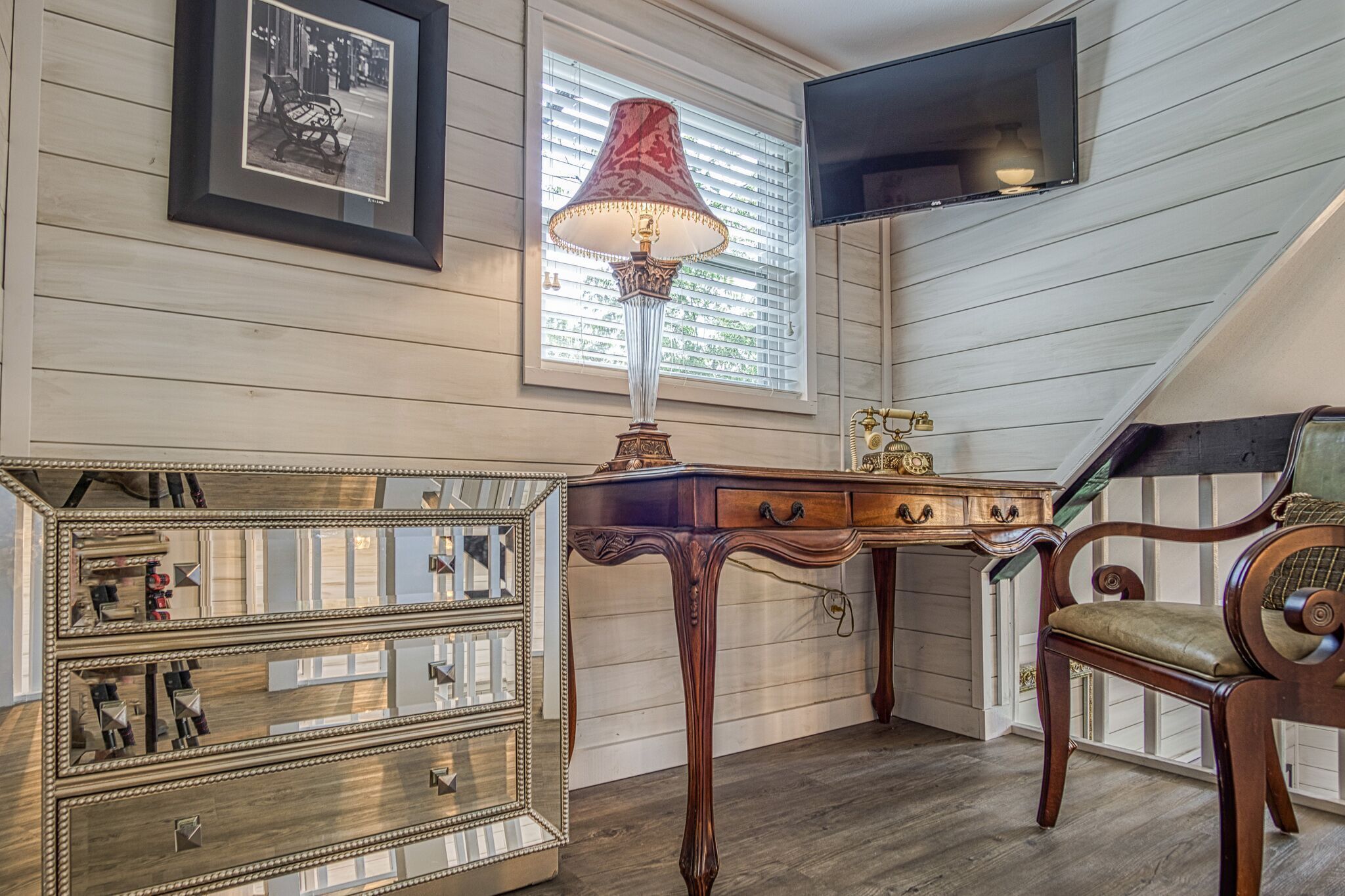 A room with a wooden desk, a chair, a mirror, a lamp, and a television mounted on the wall.