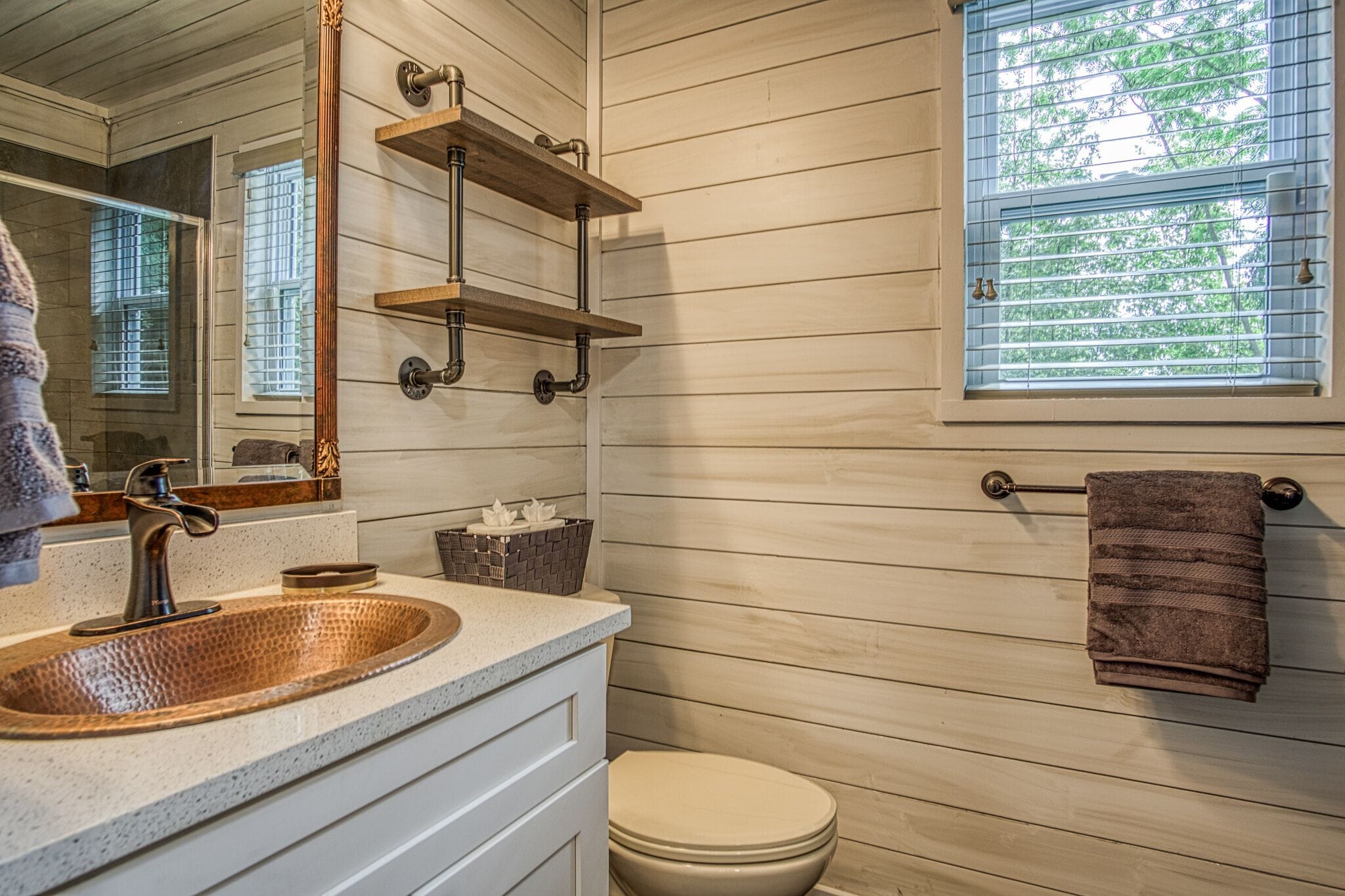 Un baño con pared de madera, un lavamanos de cobre, un espejo, un perchero para toallas y una ventana con persianas.