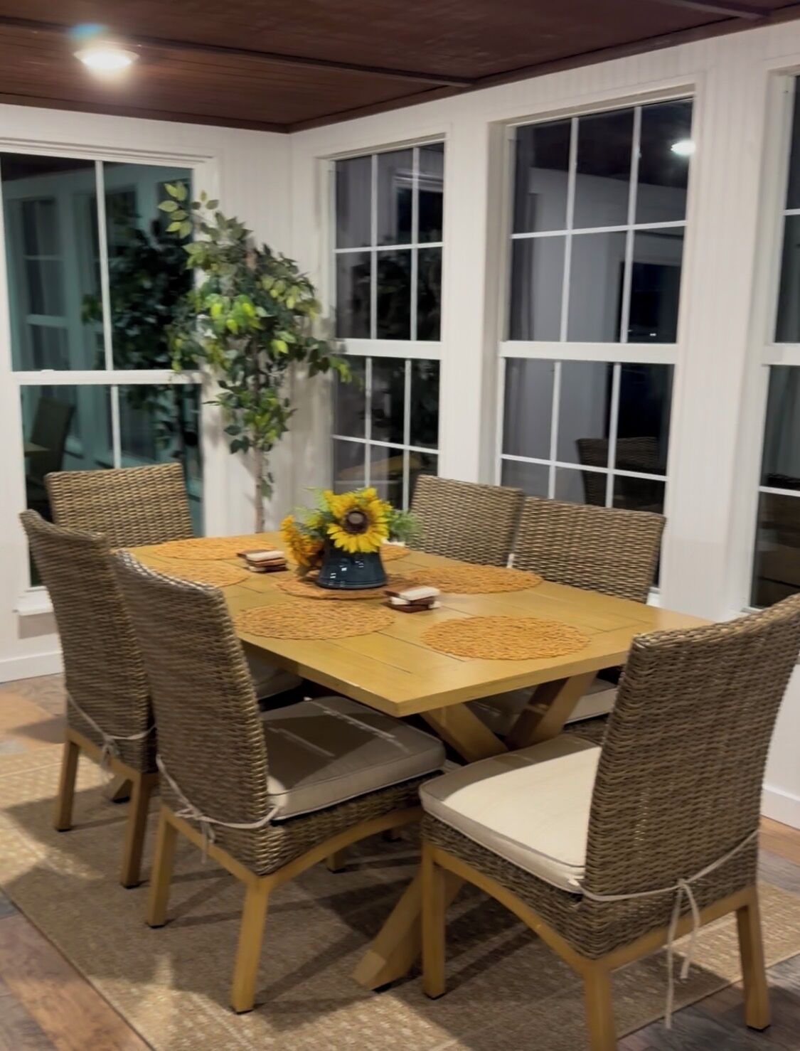 A dining area with a wooden table and wicker chairs, surrounded by large windows.