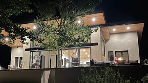 A modern house with a large tree in front, a balcony with seating, and a well-lit interior.