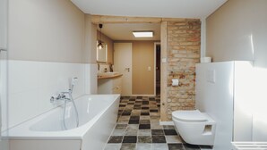 Une salle de bain avec une baignoire, des toilettes et un lavabo. Une porte en bois donne accès à une autre pièce. Le sol est recouvert de carreaux disposés en motif à carreaux.