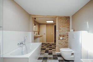 A bathroom with a bathtub, toilet, and a sink. There is a wooden door leading to another room. The floor has a checkered tile pattern.